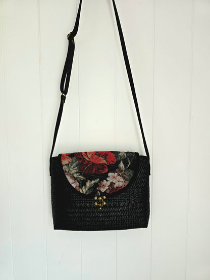 Black woven handbag with floral tapestry design on a white wooden background