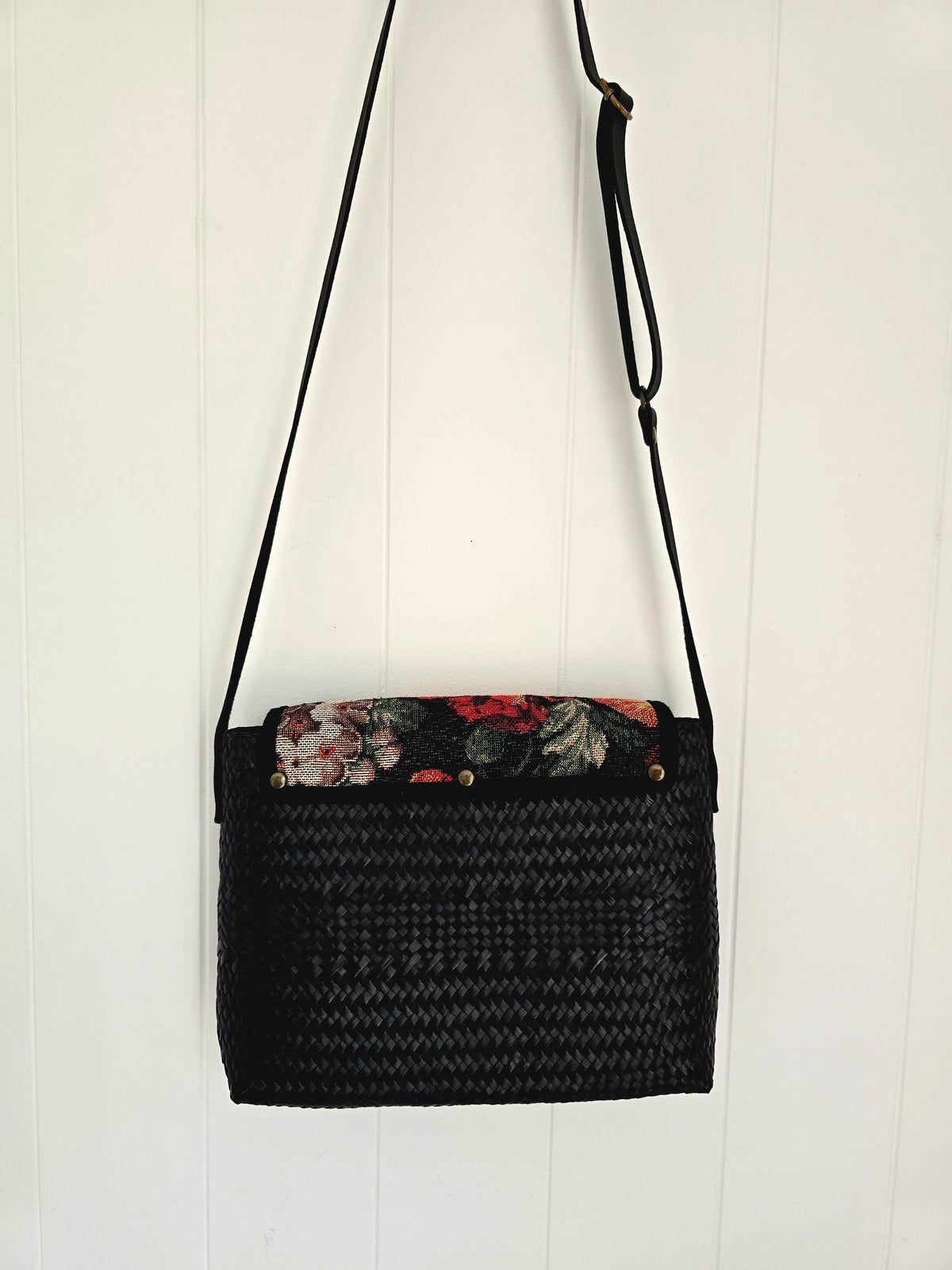 A black woven straw shoulder bag with a floral-patterned tapestry fabric lining on the top flap, hanging against a white vertical wood-paneled wall.