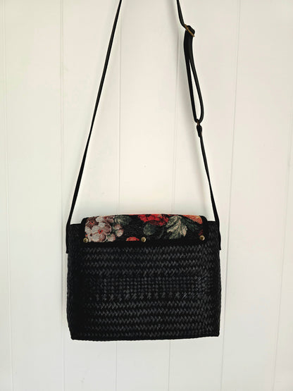 A black woven straw shoulder bag with a floral-patterned tapestry fabric lining on the top flap, hanging against a white vertical wood-paneled wall.