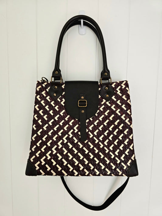 Brown and White woven handbag with faux leather flap, handles and strap against a white wall