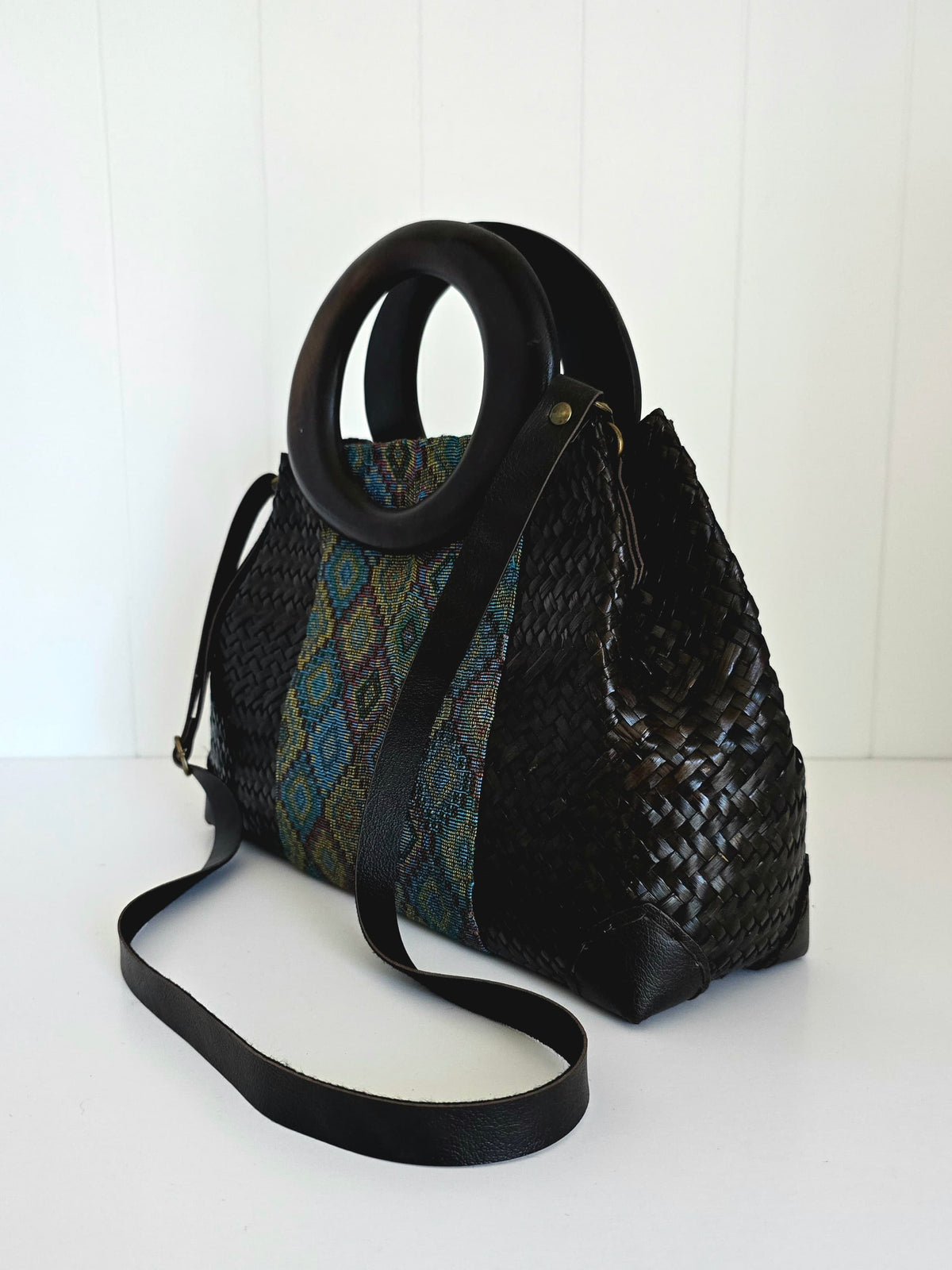 A black woven handbag with a circular black handle and a long shoulder strap, featuring a patterned textile panel on one side, photographed against a white paneled background.