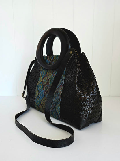 A black woven handbag with a circular black handle and a long shoulder strap, featuring a patterned textile panel on one side, photographed against a white paneled background.