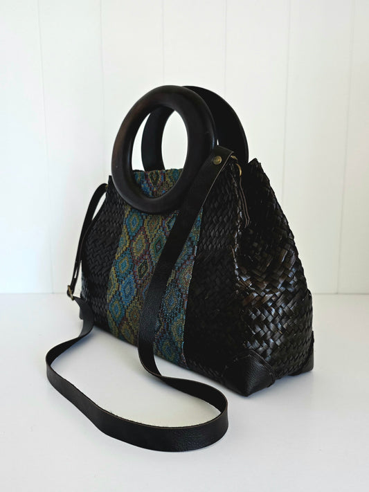 A black woven handbag with a circular black handle and a long shoulder strap, featuring a patterned textile panel on one side, photographed against a white paneled background.