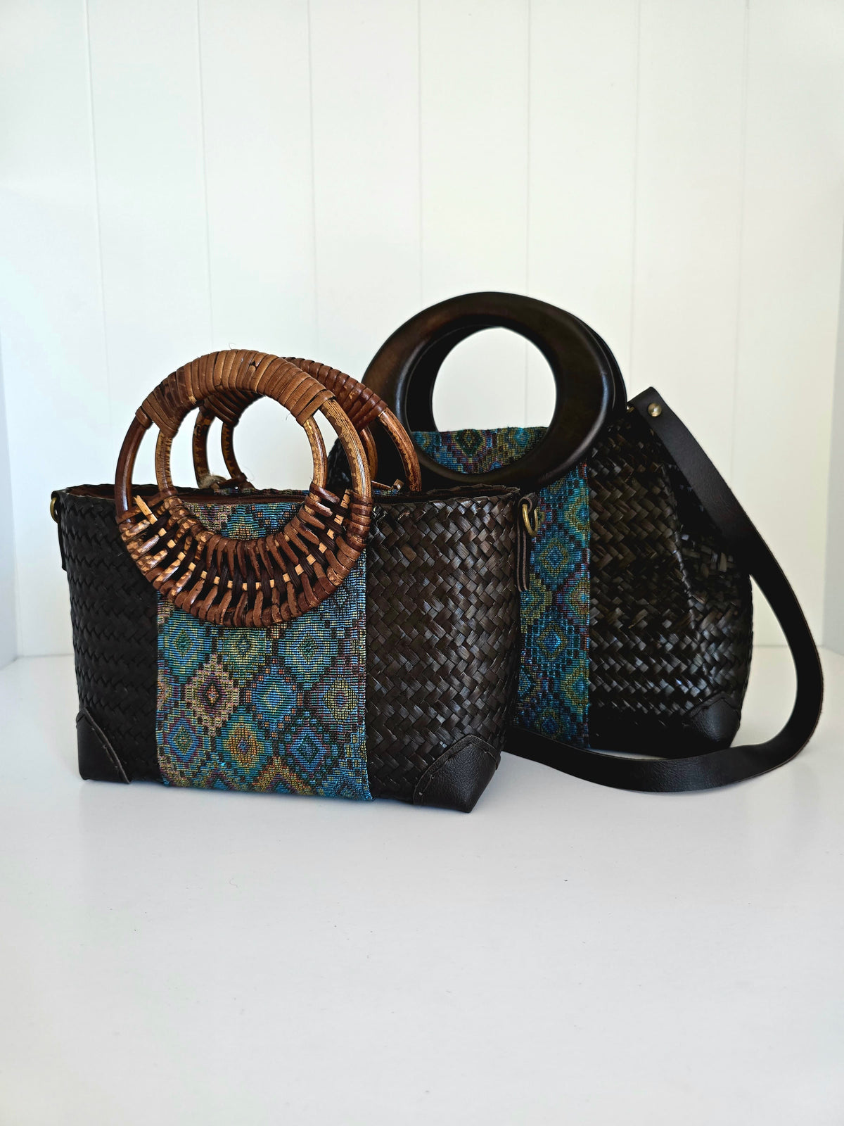 Two woven black handbags with circular brown handles and a blue geometric patterned panel, displayed against a white paneled background.