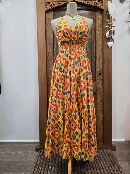 A sleeveless halter-neck maxi dress with a vibrant multicolor ikat pattern in yellow, orange, red, and blue, displayed on a mannequin in front of a rustic wooden door.