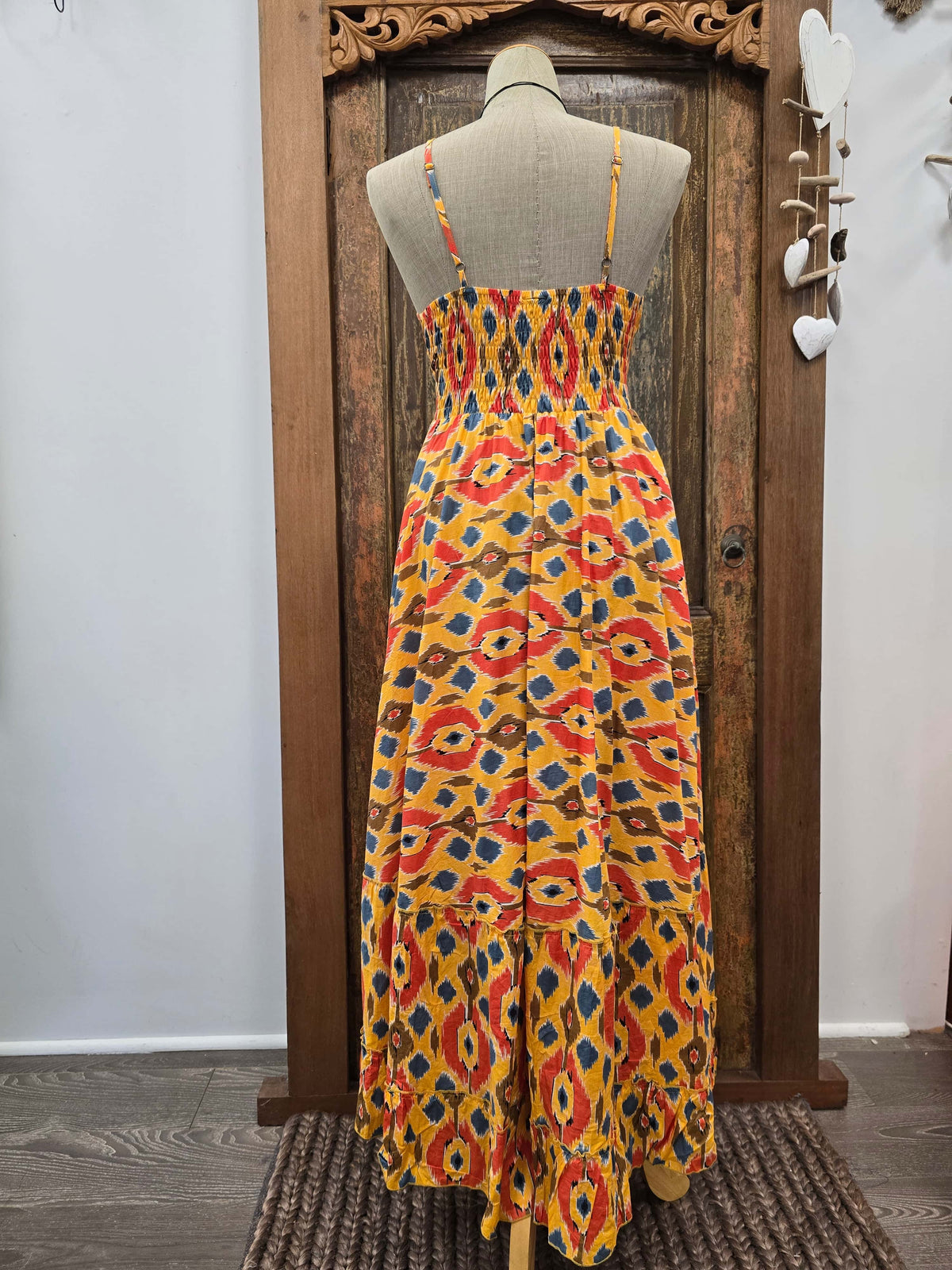A sleeveless midi dress with thin straps and a smocked bodice, featuring a vibrant yellow, red, and blue ikat pattern, displayed on a mannequin in front of a rustic wooden door.