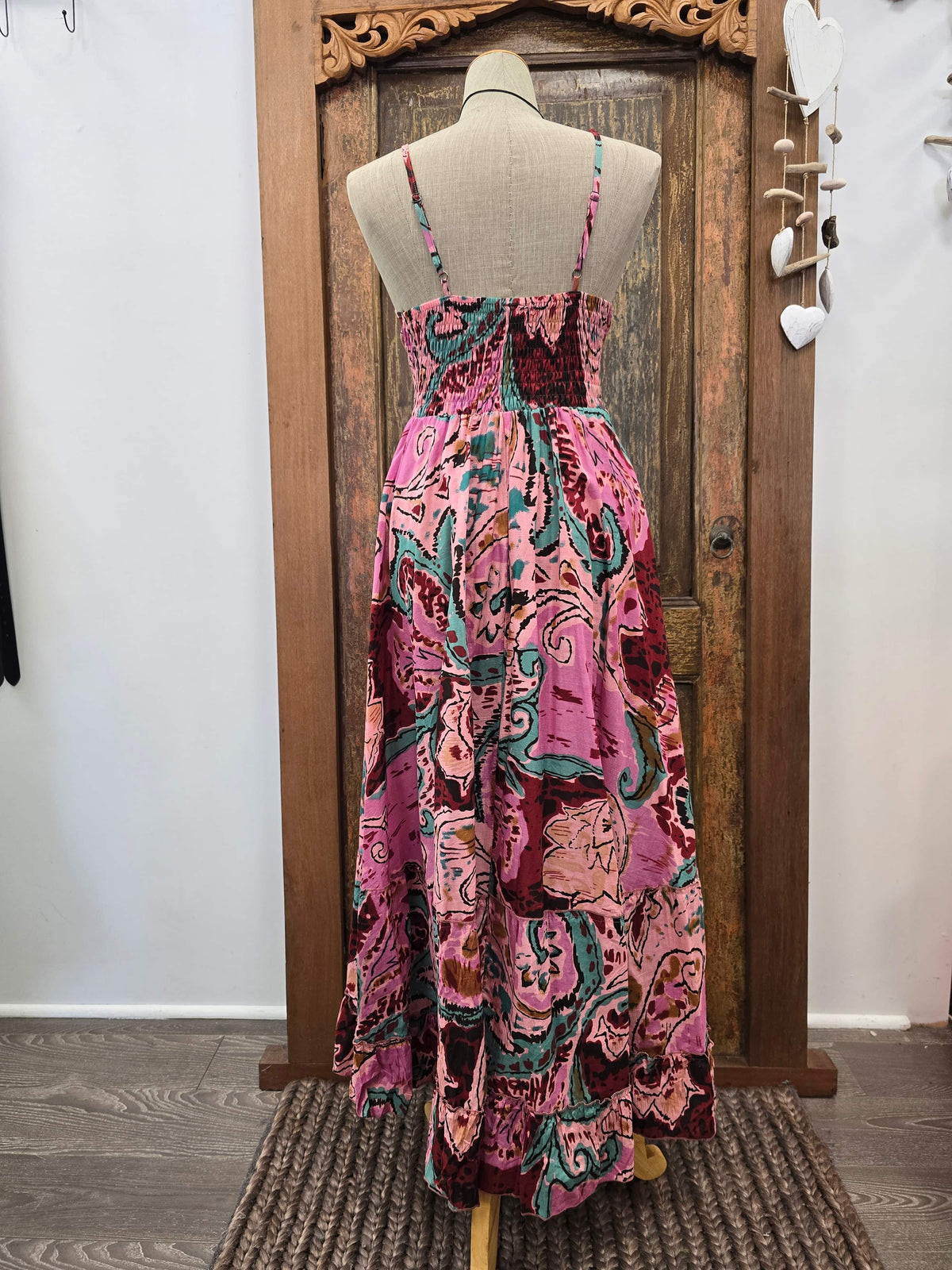 A sleeveless maxi dress with thin straps and a smocked bodice, featuring a vibrant pink, teal, and burgundy abstract print, displayed on a mannequin in front of a wooden door.