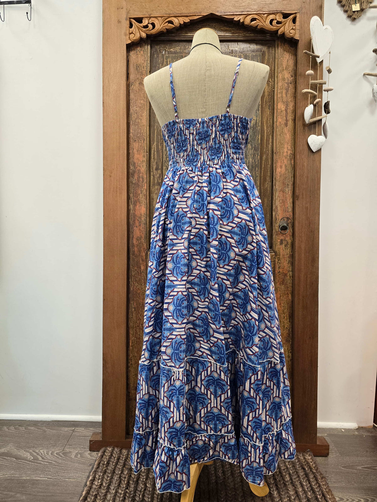 A sleeveless midi dress with a blue and white floral block print, displayed on a beige mannequin torso in front of a rustic wooden door.