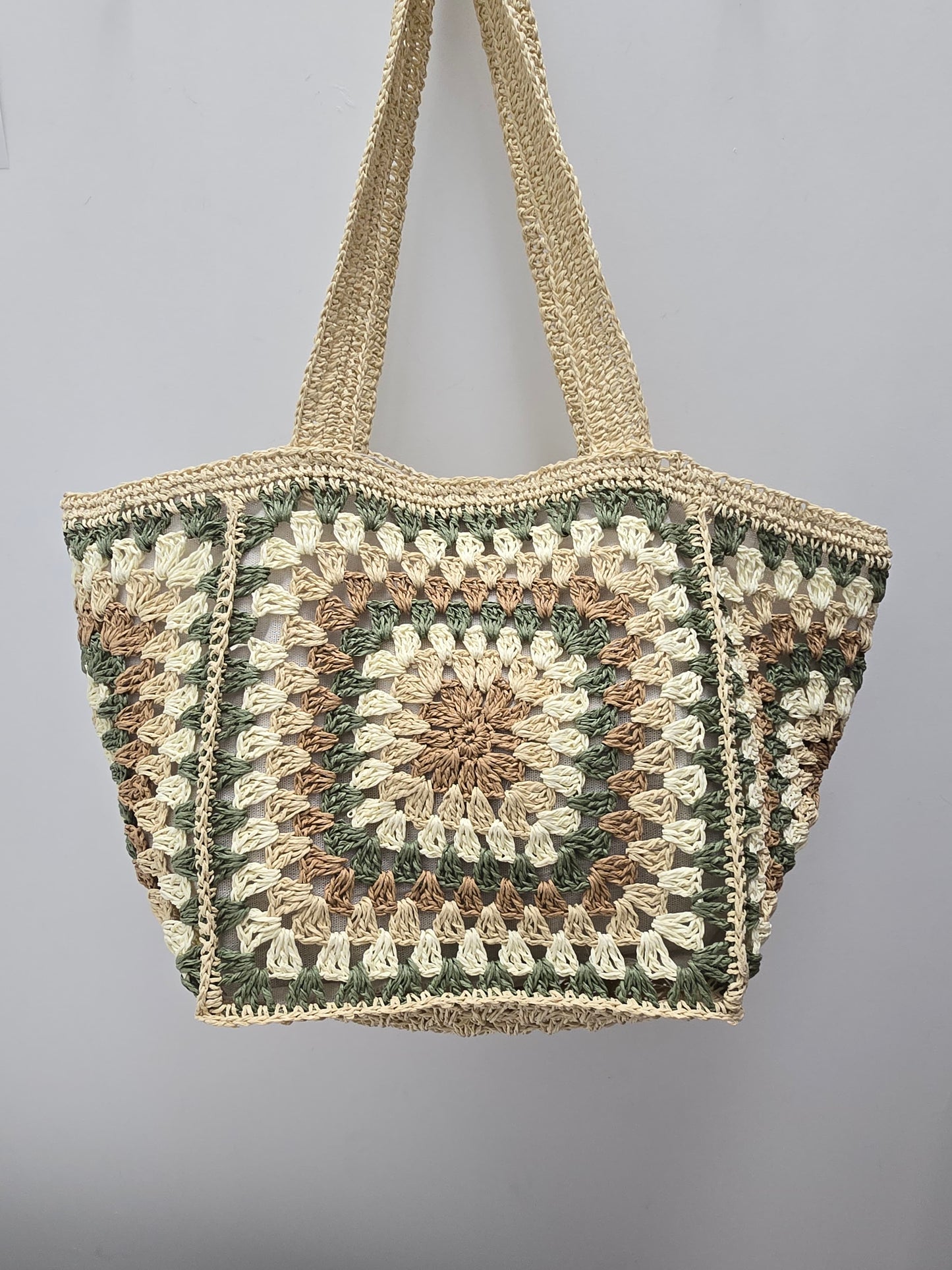 A handmade crocheted tote bag featuring a granny square pattern in natural beige, olive green, and brown, suspended against a plain light gray background.