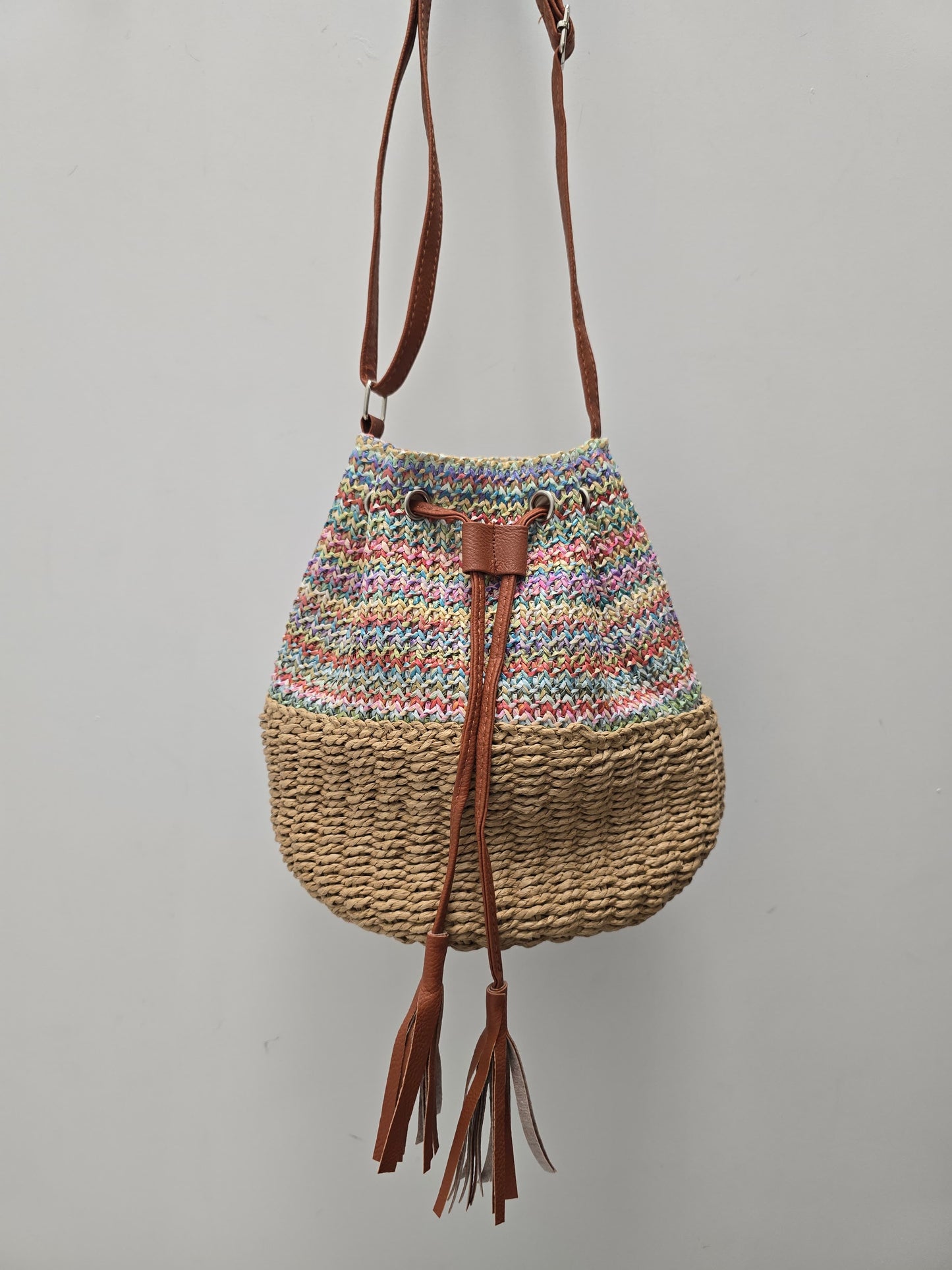 A handmade bucket bag with a multicolor striped knit top half, a natural woven straw bottom half, and brown leather tassel drawstrings, hanging against a plain gray wall.