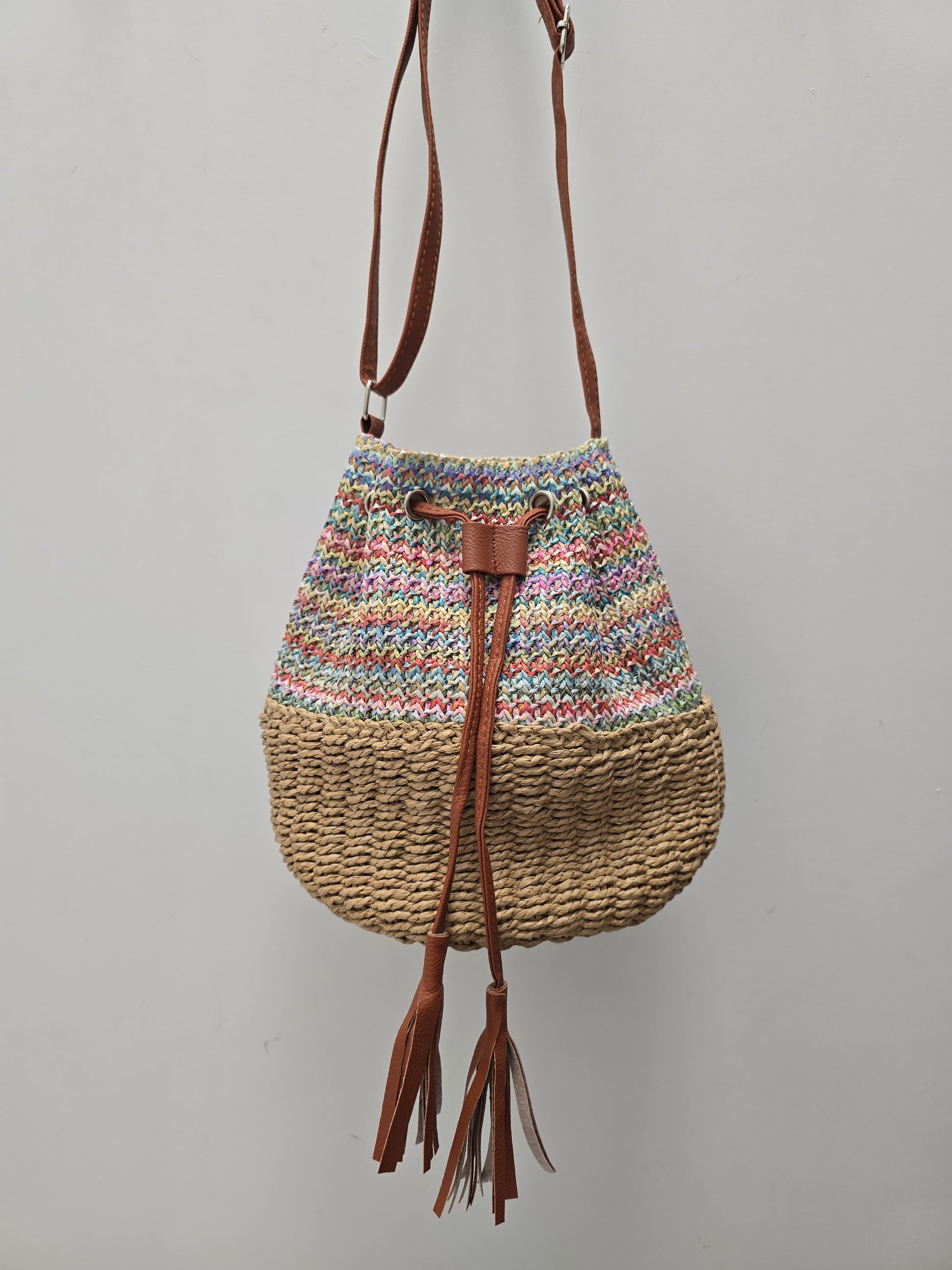 A handmade bucket bag with a multicolor striped knit top half, a natural woven straw bottom half, and brown leather tassel drawstrings, hanging against a plain gray wall.