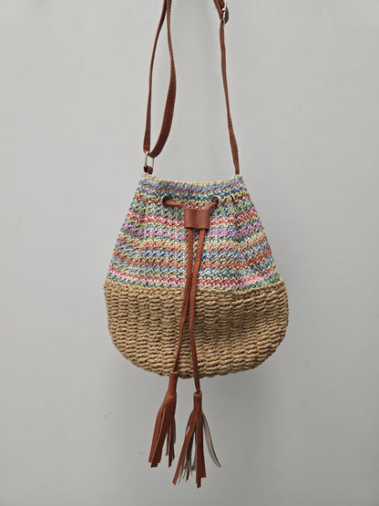 A handmade bucket bag with a multicolor striped knit top half, a natural woven straw bottom half, and brown leather tassel drawstrings, hanging against a plain gray wall.