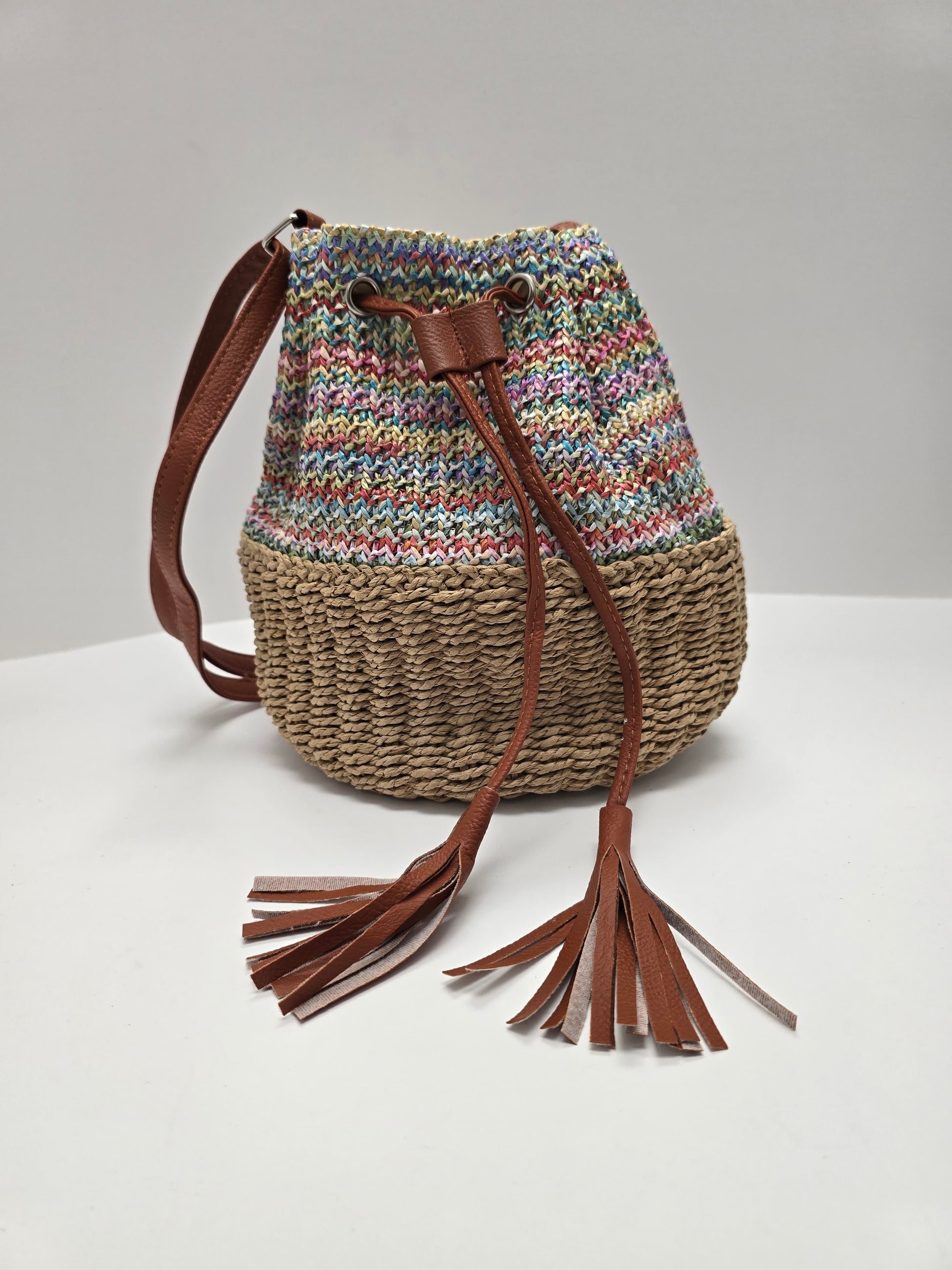 A handmade bucket bag with a multicolor knit pattern on the upper half, a natural woven straw lower half, and brown leather tassels and shoulder strap, set against a plain light gray background.