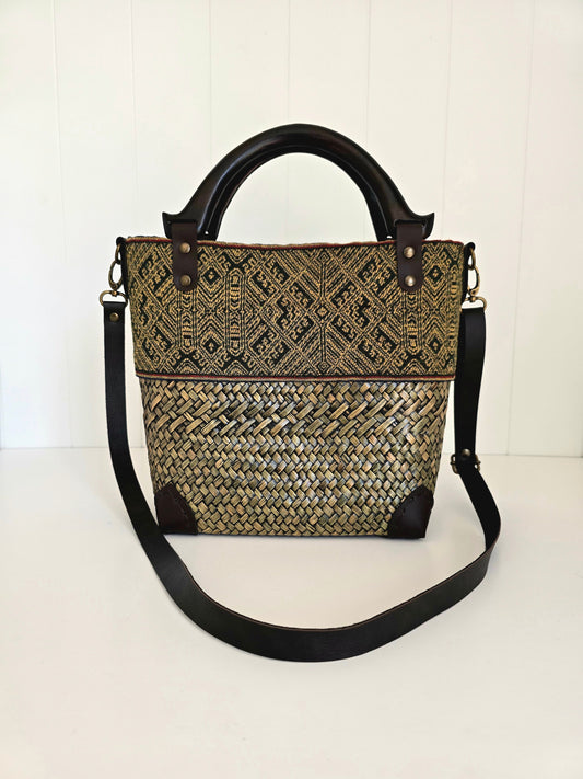 Woven natural handbag with black strap, wooden handles and natural toned material panel on a white background