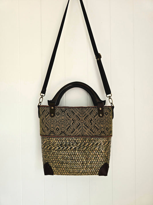 A woven tote bag with a black faux leather strap and wooden top handle, featuring a geometric patterned upper half and a textured woven lower half, hanging against a white paneled wall.