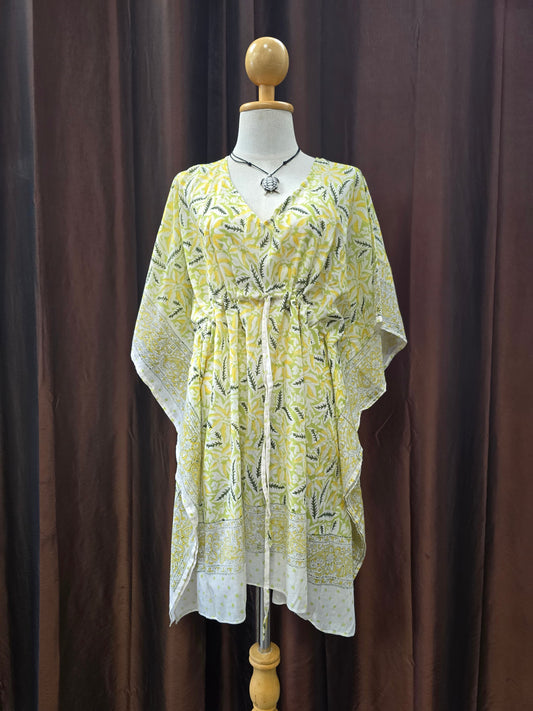 A yellow and white patterned kaftan tunic dress displayed on a wooden dress form mannequin against a dark brown curtain backdrop.